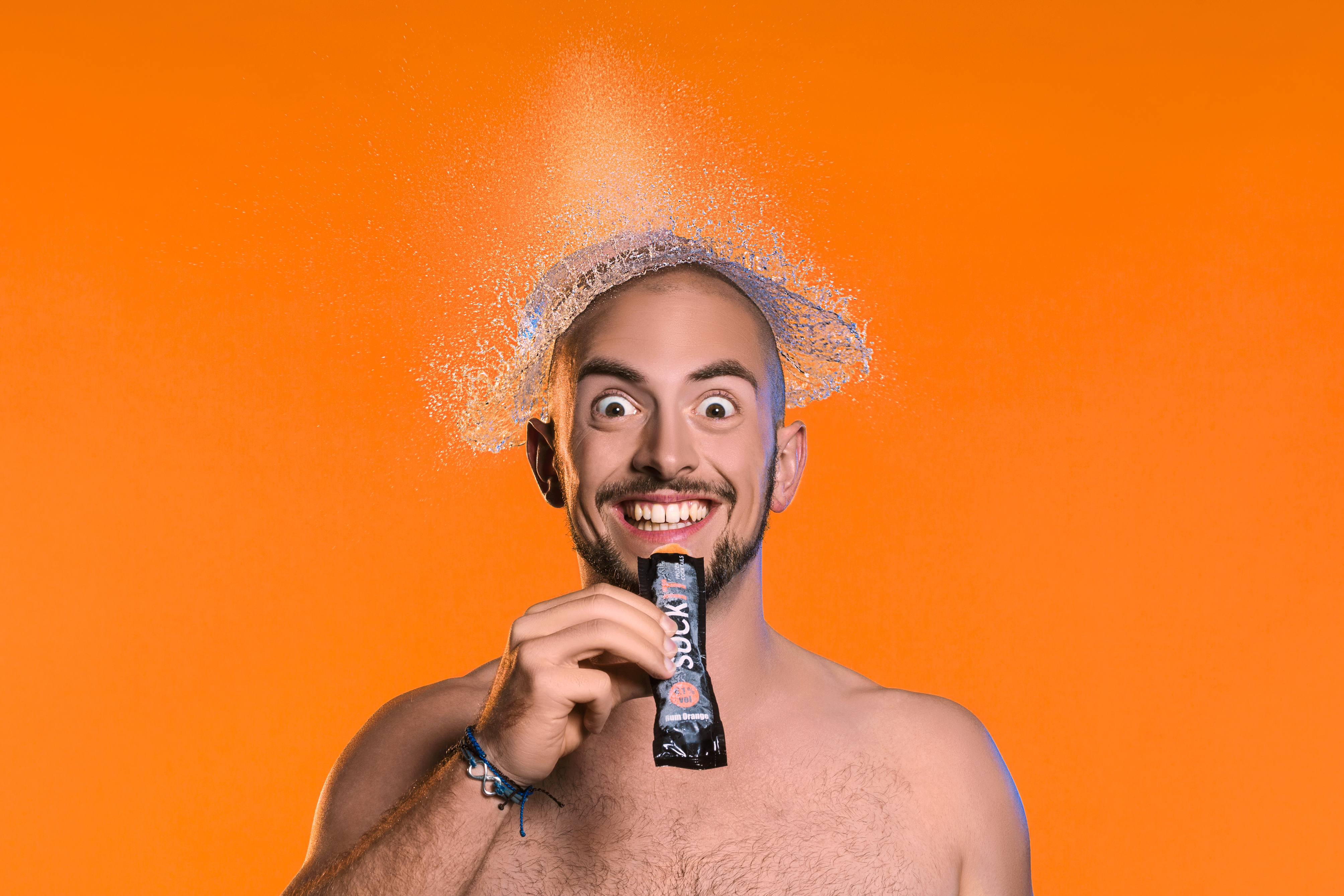 Studio-Portrait eines Mannes mit Wasserexplosion über dem Kopf vor orangenem Hintergrund. Studio portrait of a man with a water explosion over his head in front of a orange background.