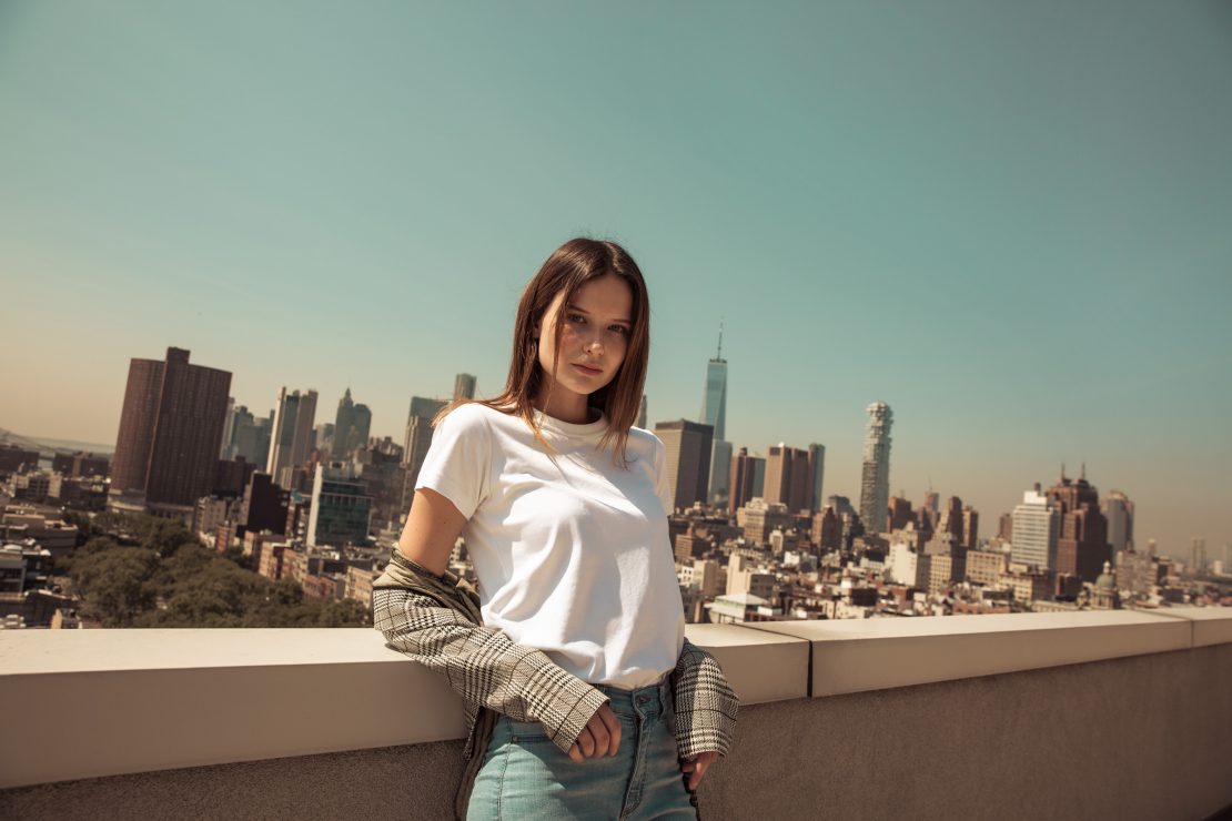 Woman in a white T-shirt and checked jacket on a rooftop overlooking the Manhattan skyline.