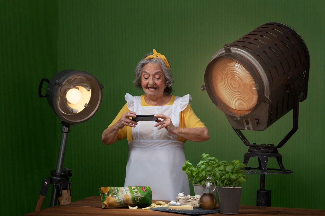 Model Luccia Guzzardi as “Nonna”, holding a bag of crisps between studio lights against a green background.